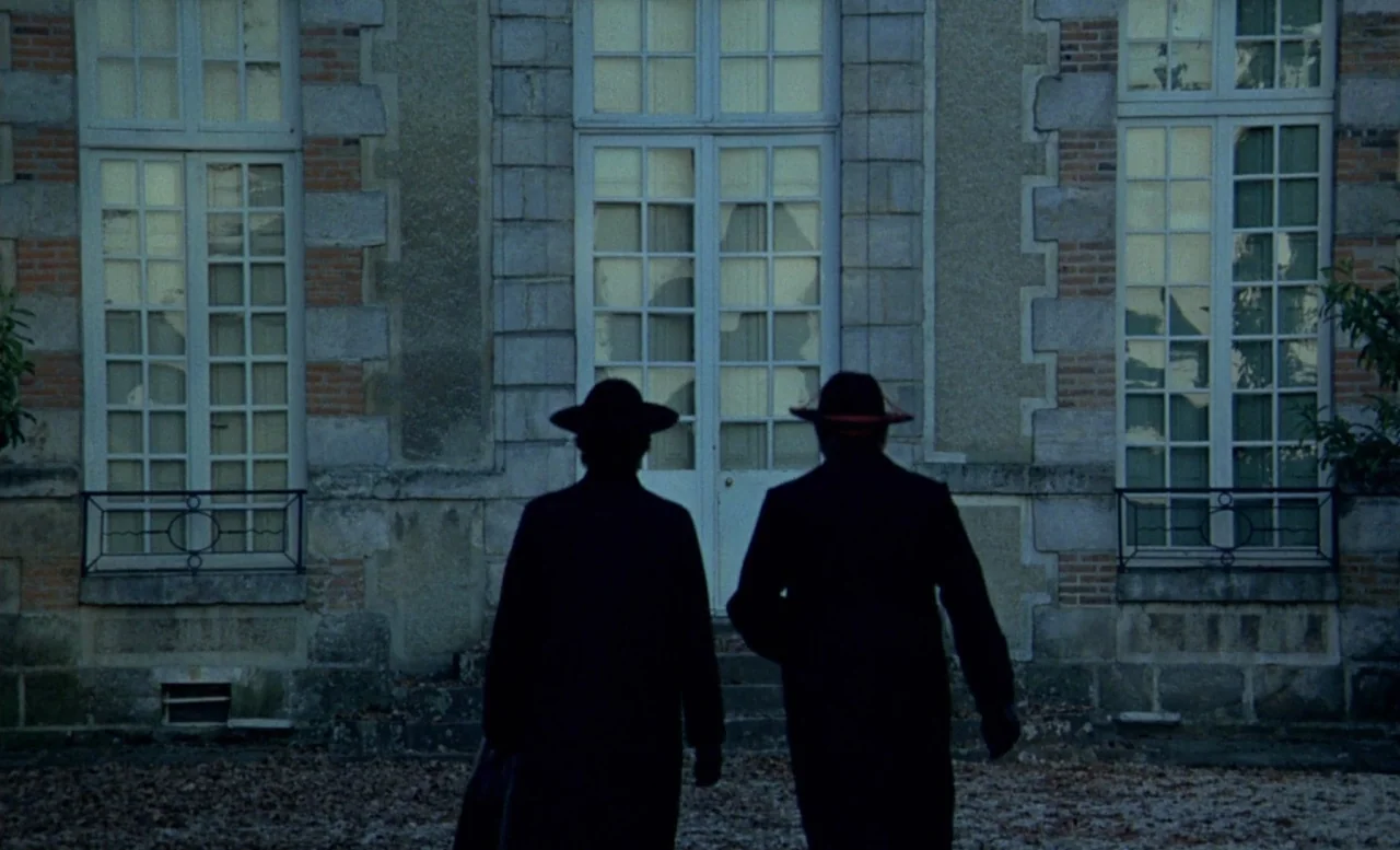 The Beast (1975) movie still: fullbody — A stone manor facade features large glass windows and arched door frames. Two men in dark coats and hats…