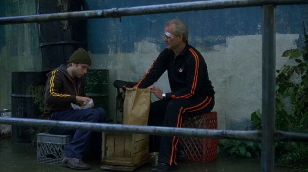 Broken Flowers movie still: medium — Two men sit on plastic crates against a weathered blue wall in an urban alley. The older man with a forehead…