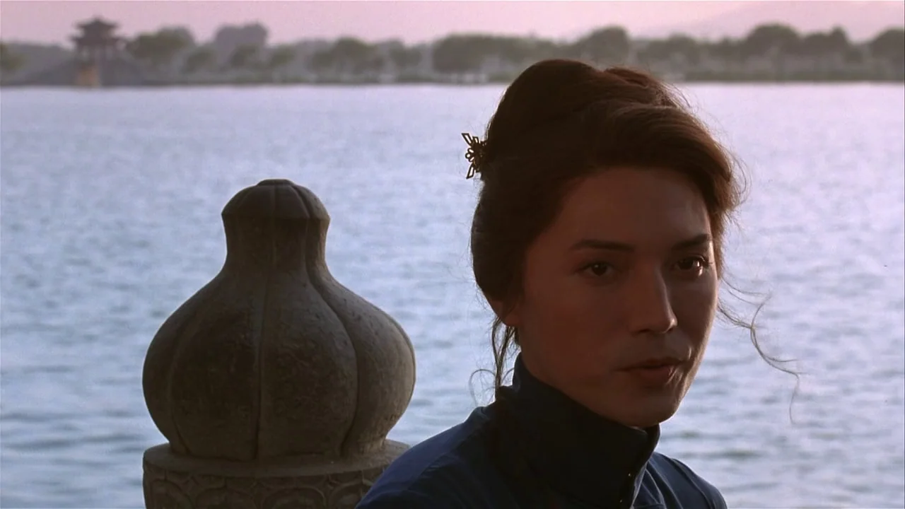M. Butterfly (1993) movie still: closeup — A person stands beside a carved stone balustrade overlooking a wide, calm lake. In the distance, a…