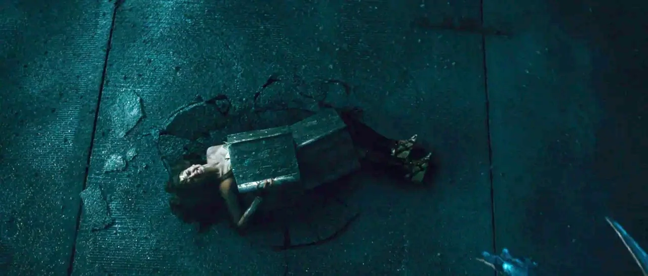 Wonder Woman (2017) movie still: wide — A dark asphalt surface features a large crater with jagged broken concrete fragments. A woman in metallic…