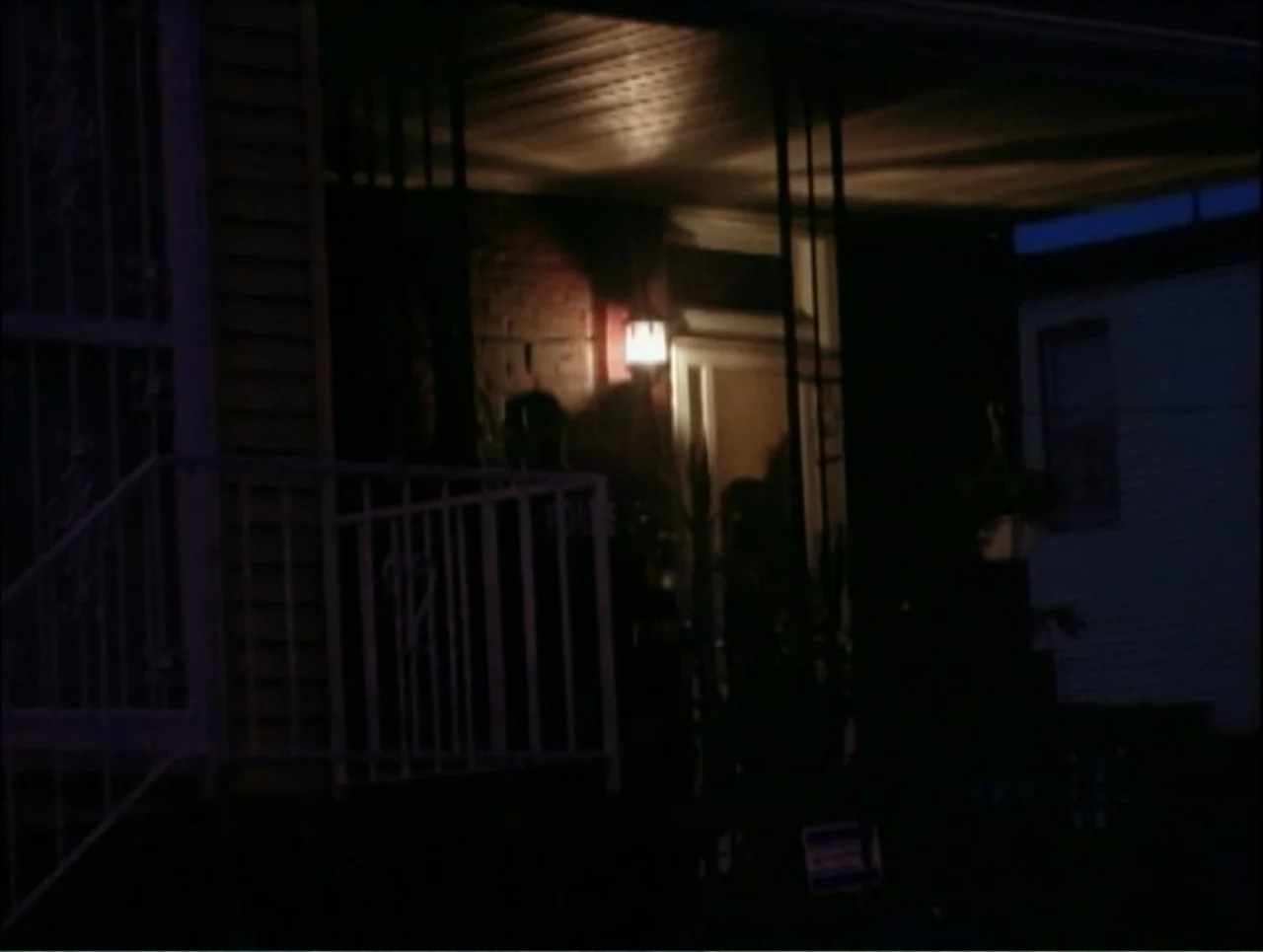 Addiction (2024) movie still: medium — A dark house porch features brick walls and a metal railing at night. A person stands partially hidden in…