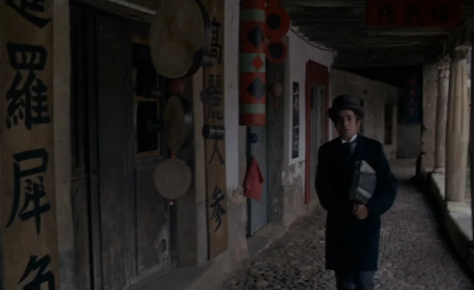 The Immortal Story (1968) movie still: fullbody — A stone-paved covered walkway with traditional Chinese signage and hanging drums on wooden posts.…