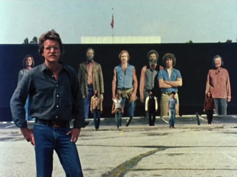 Mur Murs (1981) movie still: fullbody — An asphalt parking lot features a large black billboard displaying portraits of several people. Smaller…