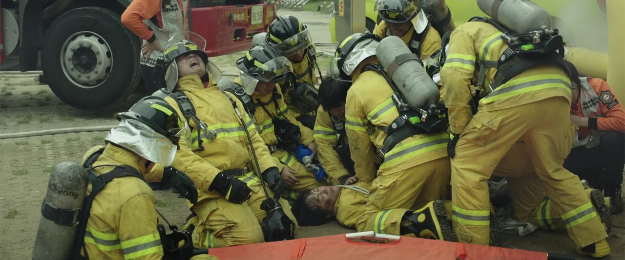 Along with the Gods: The Two Worlds (2017) movie still: medium — A group of firefighters in yellow gear cluster around a victim on the ground. They…