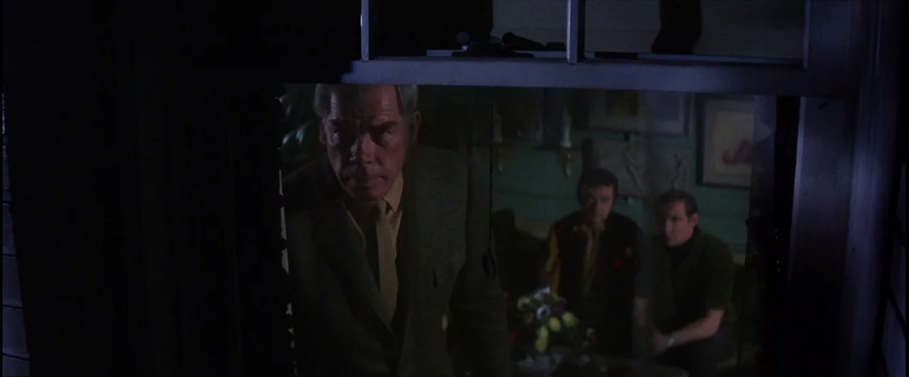 Point Blank (1967) movie still: medium — An older man looks through a pane of glass from a dark exterior space. Two men sit at a table inside a…