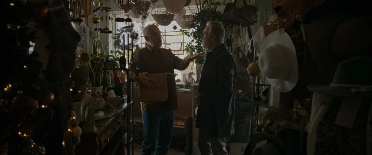 Silent Night, Deadly Night (2025) movie still: medium — A cluttered shop interior filled with hanging baskets, hats, and holiday decorations. Two…