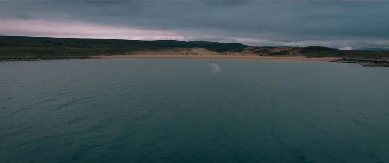 Out of Darkness (2022) movie still: establishing — An expansive coastal bay under a heavy, overcast, dark gray sky. A small boat traverses the water…
