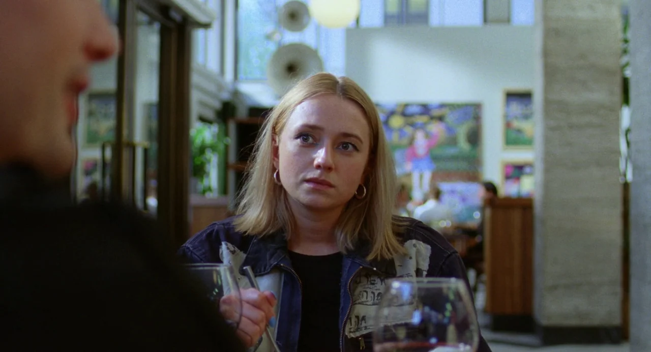 Sick of Myself (2022) movie still: medium — A restaurant interior features modern artwork on white walls and large glass panels. A woman sits at a…