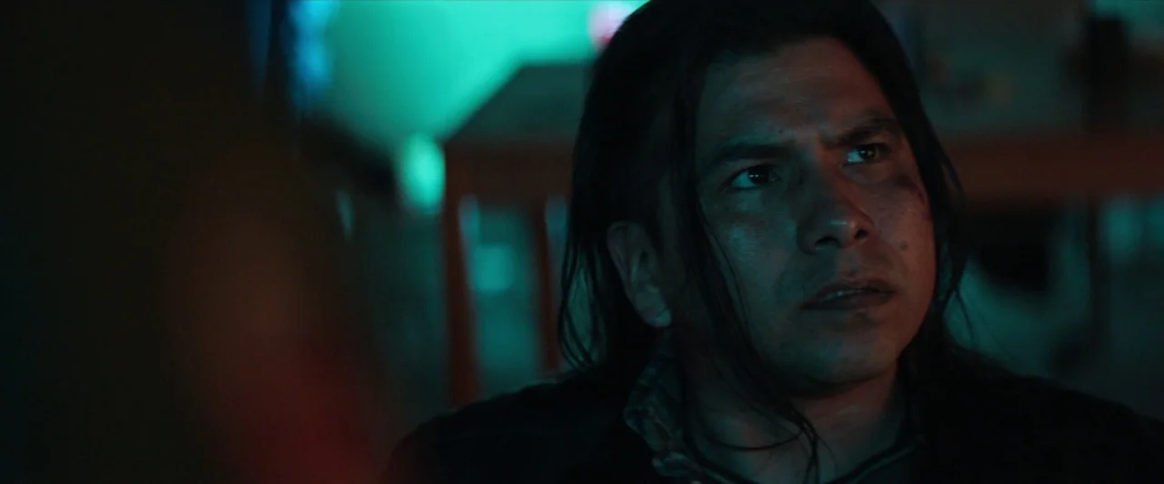 The Silencing (2020) movie still: closeup — A dimly lit indoor space is bathed in a distinct neon-green glow. A man with dark shoulder-length hair…