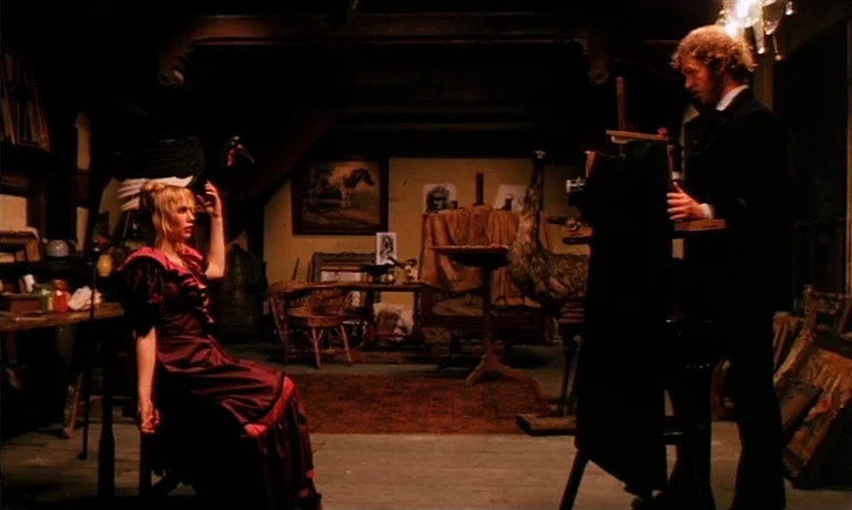Katie Tippel (1975) movie still: fullbody — An dimly lit, cluttered vintage studio contains various paintings, taxidermy, and wooden furniture…
