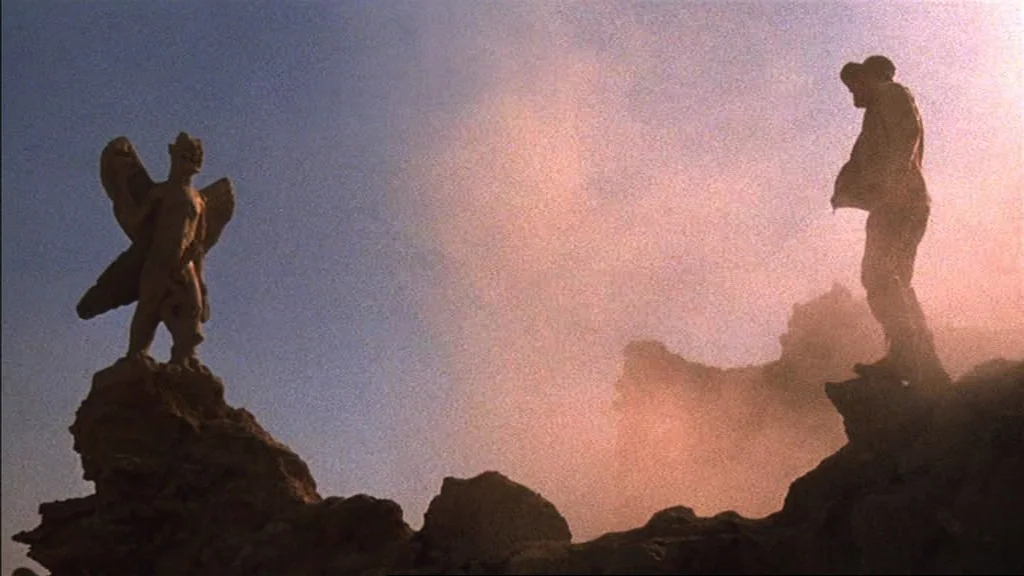 The Exorcist (1973) movie still: wide — A craggy cliffside features a stone gargoyle statue standing against a soft golden sky. A man stands on an…
