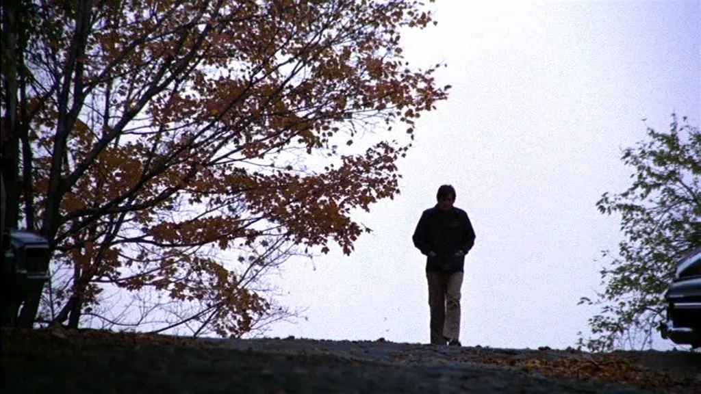 The Exorcist (1973) movie still: wide — A man walks along a dirt path lined with deciduous trees in autumn. Fallen leaves litter the foreground…