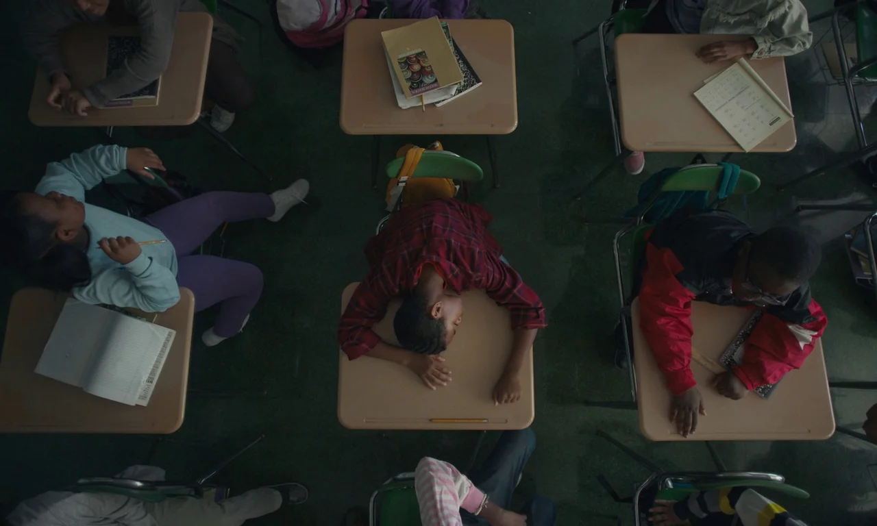 We Grown Now (2023) movie still: wide — A top-down view of multiple student desks in a school classroom. A student lies sleeping with their head…