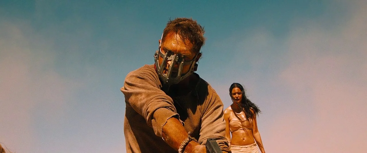 Mad Max: Fury Road (2015) movie still: medium — A vast, dusty desert expanse under a clear, bright sky serves as the backdrop. A man wearing a…