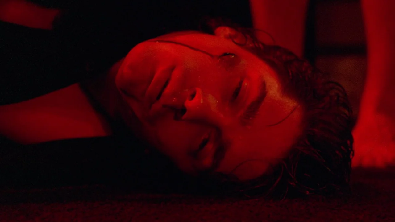 Society (1989) movie still: closeup — A man lies on a floor in a dark, dimly lit environment. He remains motionless under a wash of intense red light.