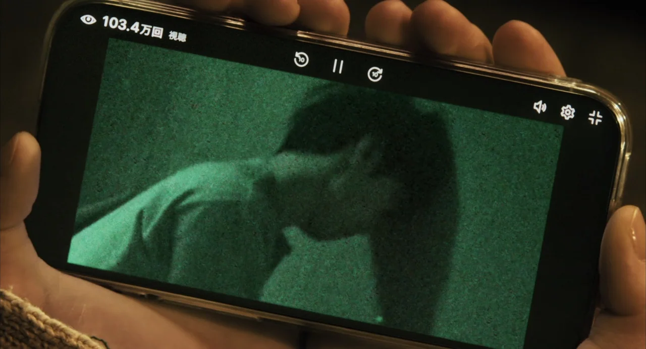 Stigmatized Properties: Possession (2025) movie still: closeup — A pair of hands holds a smartphone displaying a grainy green-tinted video feed. The…