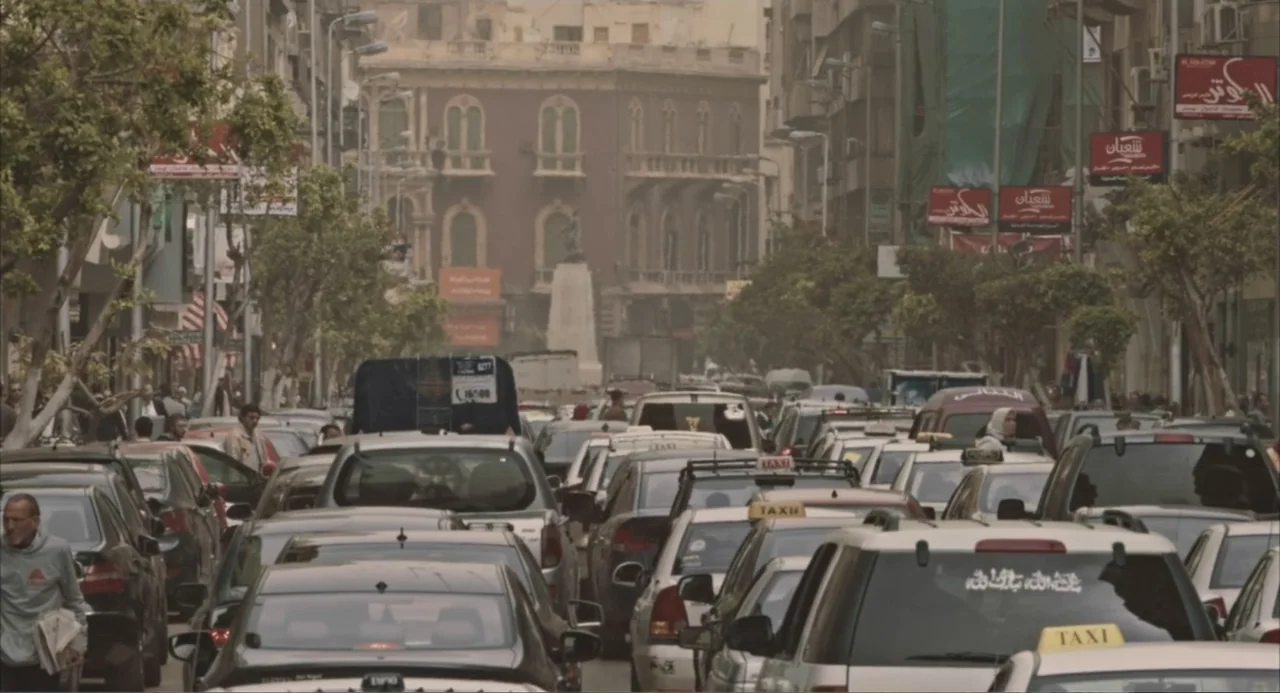 Cairo Jazzman (2017) movie still: wide — A dense urban city street is packed with numerous cars and taxis. The hazy environment includes multi-story…