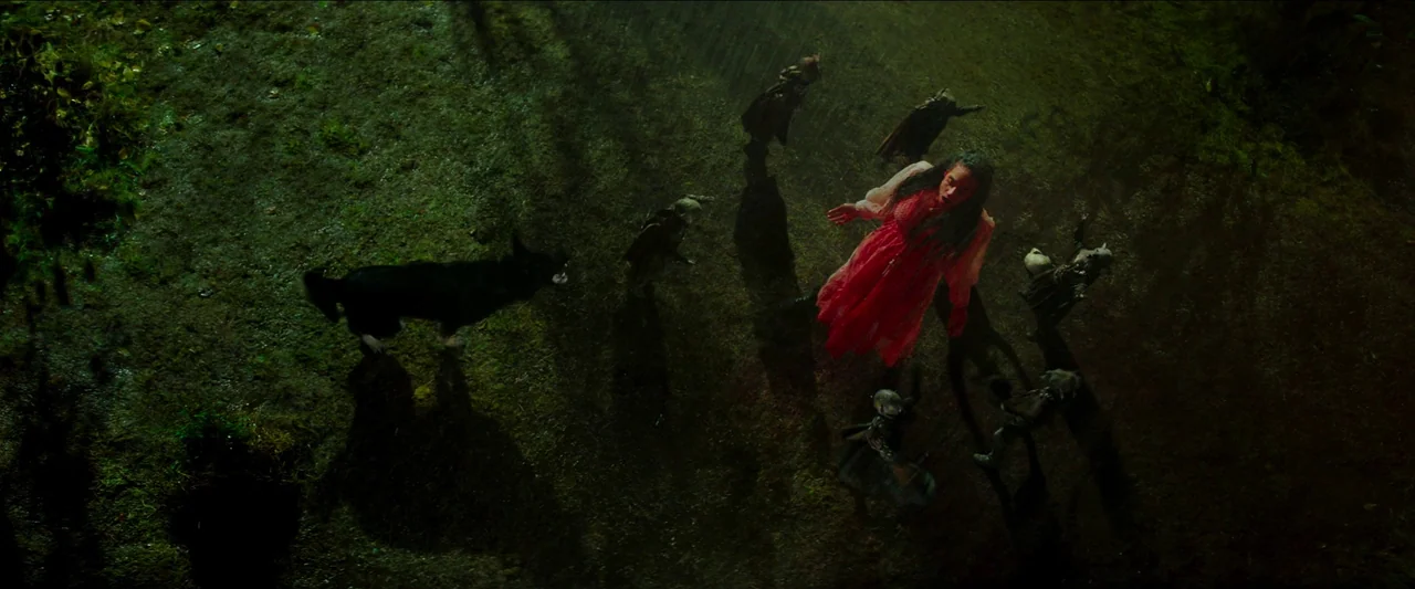 Unwelcome (2022) movie still: wide — A dark grassy area at night is illuminated by harsh lighting from above. A woman in a red dress is surrounded…