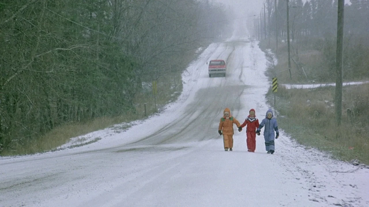 The Brood (1979) movie still: wide — A snowy rural road is flanked by bare trees and telephone poles under fog. Three children in colorful winter…
