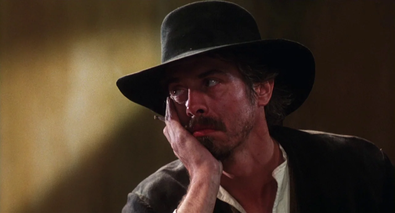 Caravaggio (1986) movie still: closeup — A dark indoor setting with a plain background suggesting a historical era. A man wearing a cowboy hat leans…