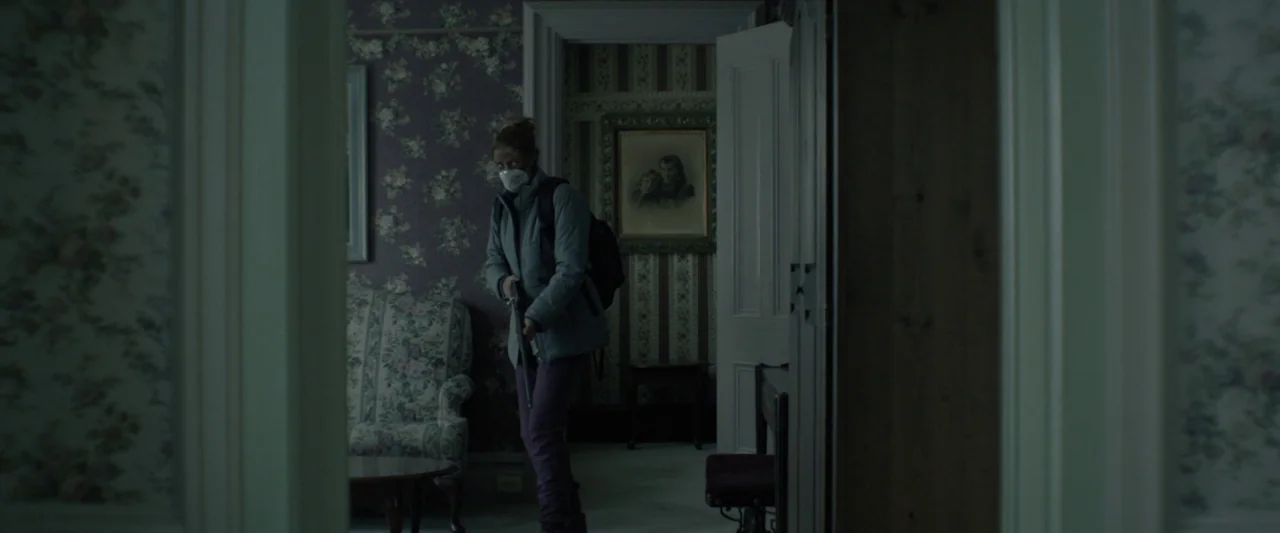 Here Alone movie still: medium — A dark house interior features floral patterned wallpaper and framed artwork on the walls. A woman wears a mask and…