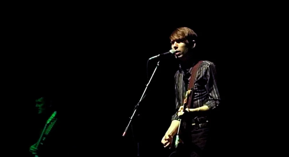 9 Songs (2004) movie still: medium — A dimly lit musical performance space with total darkness surrounding the center subject. A man plays guitar…
