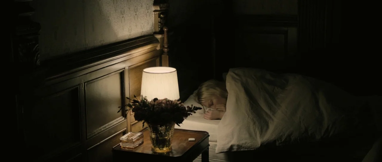 Melancholia (2011) movie still: medium — A dimly lit room features a carved wooden headboard against a patterned wallpaper background. A blonde…