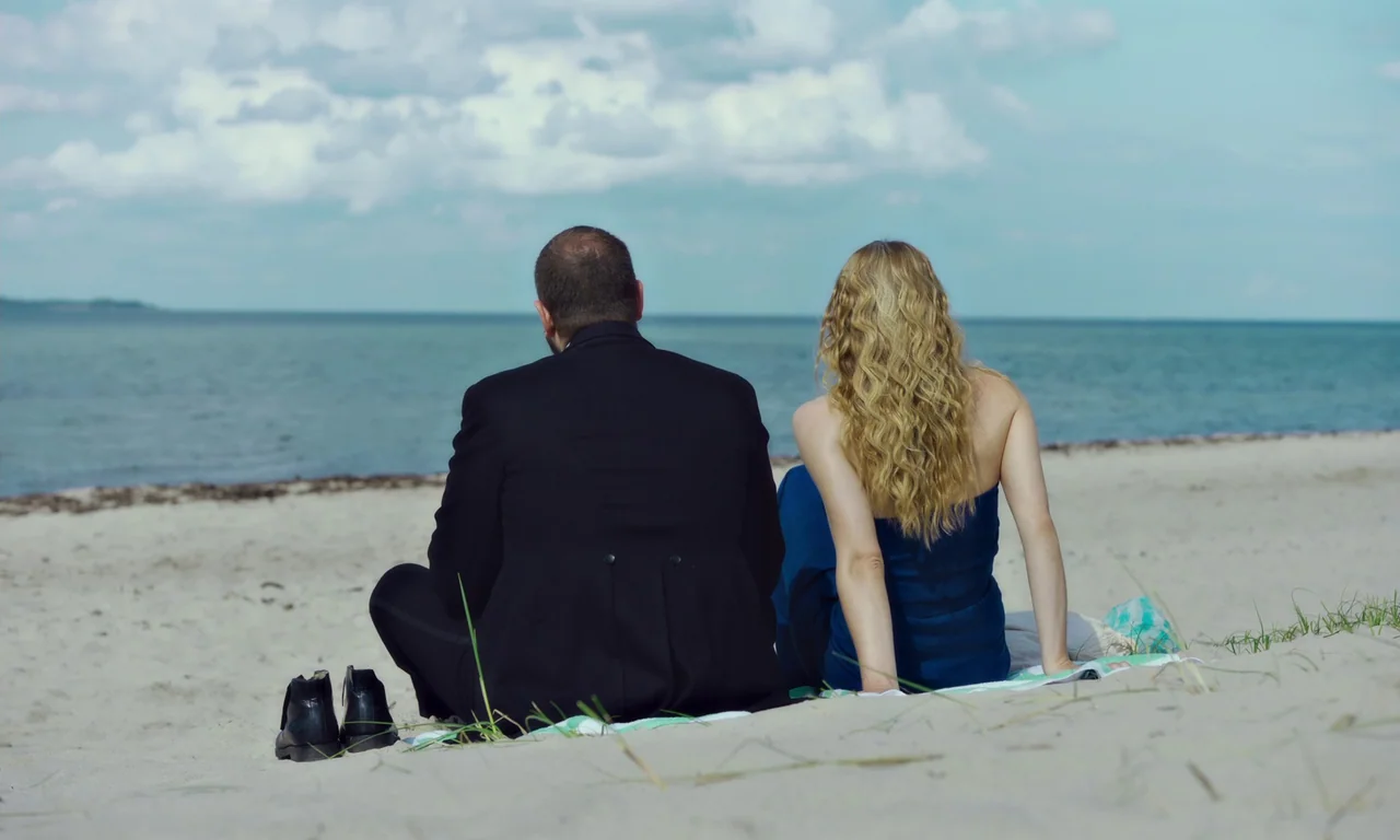 Bloodsuckers (2021) movie still: fullbody — A man in a tuxedo and a woman in a blue dress sit on a beach. They face toward the calm water and…