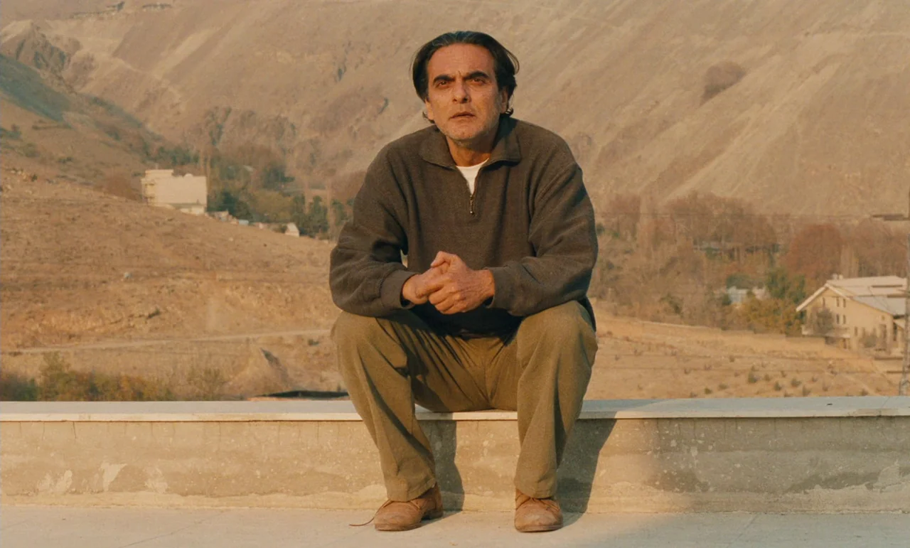 Taste of Cherry (1997) movie still: medium — A man sits on a concrete ledge against a dry, mountainous desert landscape backdrop. The setting…