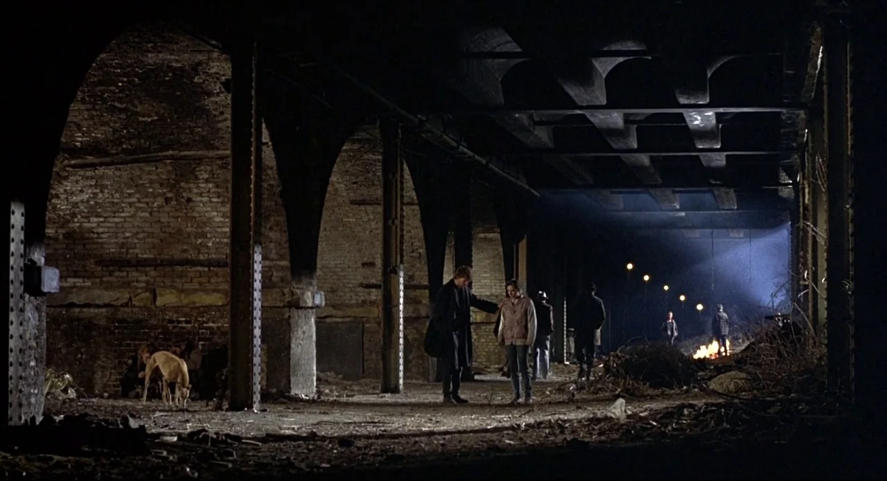 Naked (1993) movie still: wide — A dark concrete underpass with weathered brick walls and heavy support pillars. Several figures gather in the…