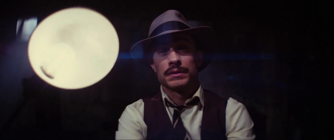 Neruda (2016) movie still: medium — A dark space features a man lit by an intense nearby lamp. The man stares ahead while wearing a vintage fedora…