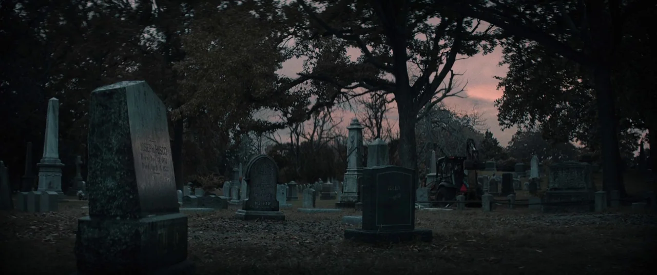 The Many Saints of Newark (2021) movie still: wide — A quiet cemetery at dusk with numerous weathered stone monuments and leafless trees. A…