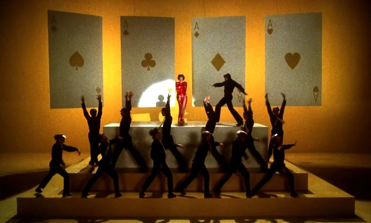 New York, New York (1977) movie still: wide — A theatrical stage features four giant oversized playing cards against a vibrant yellow backdrop. A…