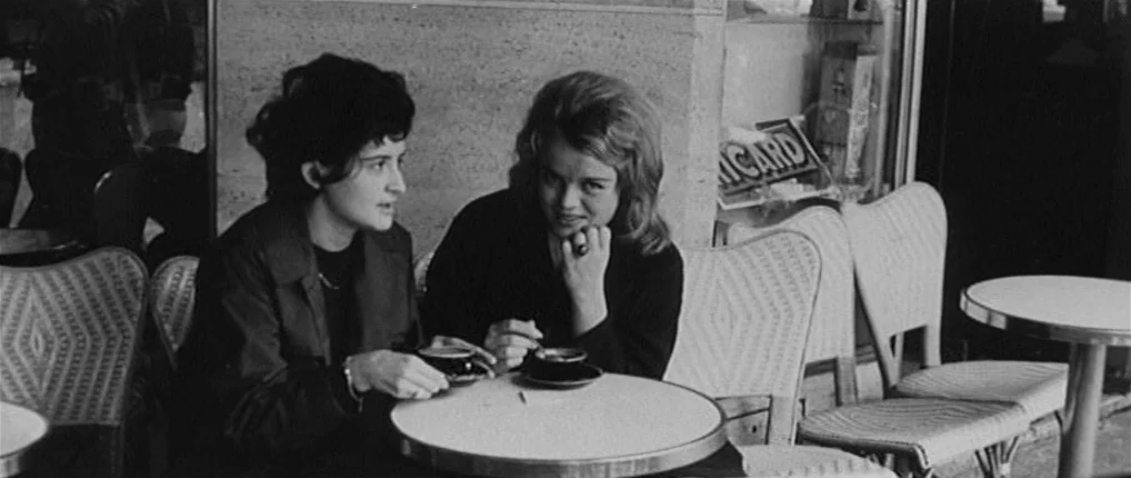 Antoine and Colette (1962) movie still: medium — A sidewalk cafe features woven chairs and a small round metal-edged table. Two young women sit…