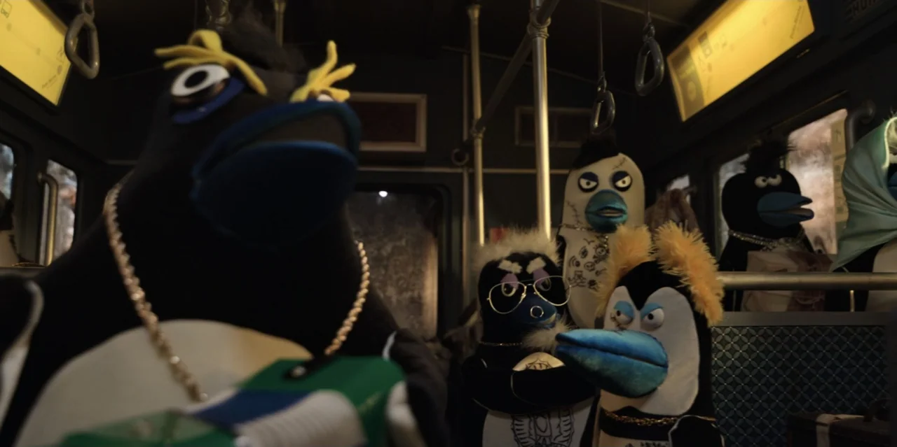 31 Minutes: One Hot Christmas (2025) movie still: medium — A dark interior of a public bus filled with anthropomorphic penguin puppets. Several…