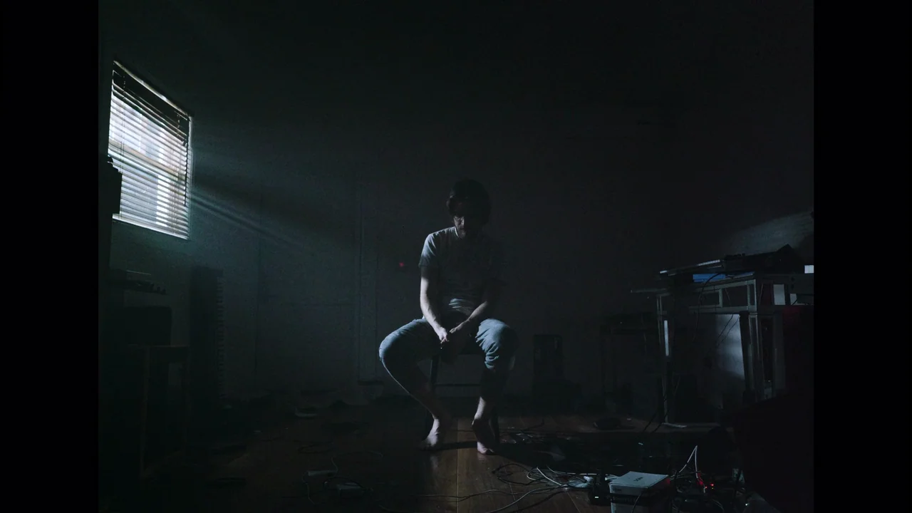 Bo Burnham: Inside (2021) movie still: fullbody — A dark, sparsely furnished room contains a single man sitting on a wooden chair. Light streams…