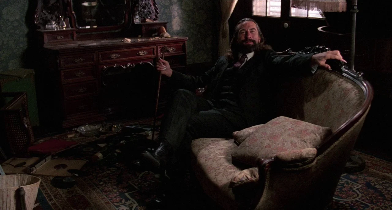 Angel Heart (1987) movie still: medium — A cluttered room features a dark wood dressing table and a vintage upholstered sofa. A bearded man in a…