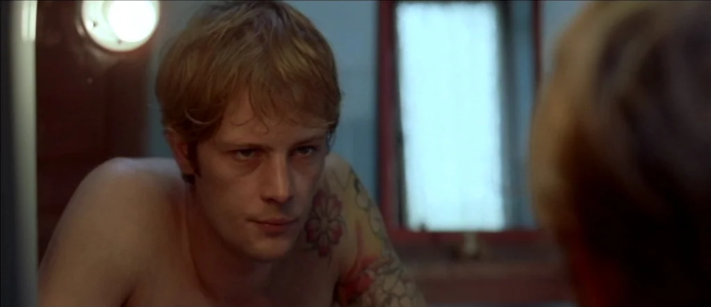 White Material (2009) movie still: closeup — A small room features a mirror reflecting a man with tattoos on his shoulder. The man stares directly…