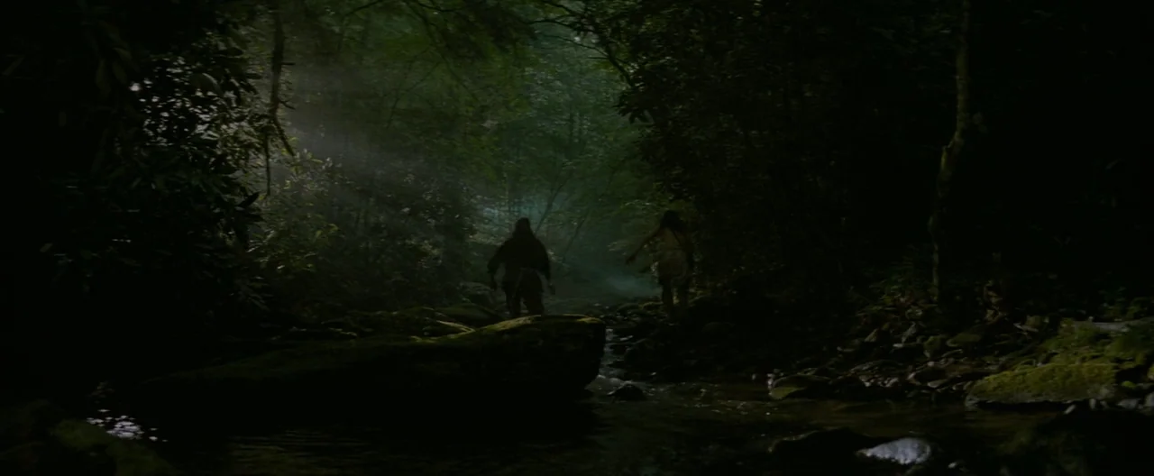 The Last of the Mohicans (1992) movie still: wide — A dark, dense forest contains a shallow rocky stream with thick overhead canopy cover. Two…