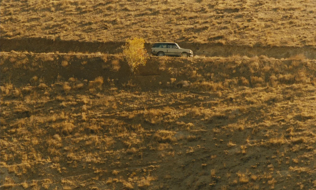Taste of Cherry (1997) movie still: wide — An arid, grassy hillside features a winding dirt track cut into its slope. A light-colored four-door…
