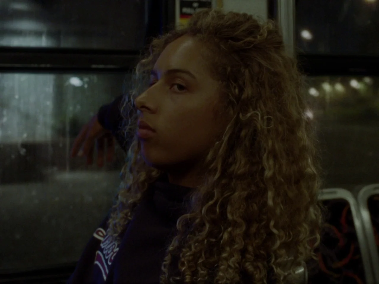 Mid90s (2018) movie still: medium — A dimly lit public transport vehicle at night with rows of patterned seating visible. A young woman with curly…