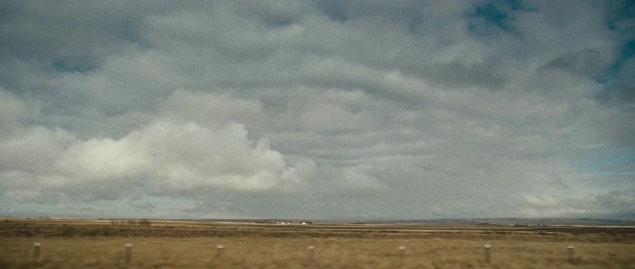 Surveillance (2008) movie still: establishing — A vast, flat, dry grassy expanse extends beneath a heavy, cloud-filled overcast sky. A line of low…