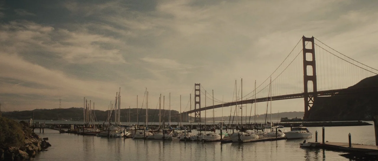 The Diary of a Teenage Girl (2015) movie still: establishing — A marina with docked sailboats sits in the foreground below a suspension bridge. The…