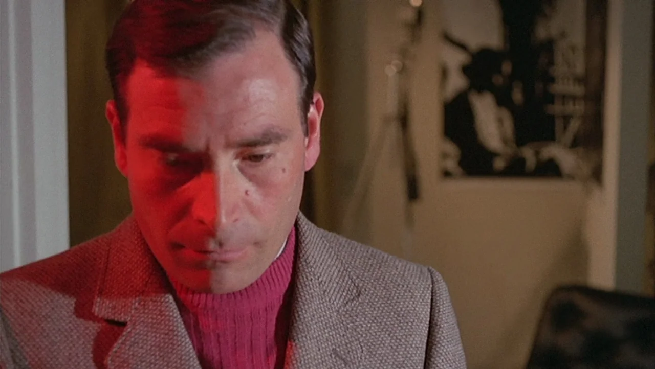 The Black Belly of the Tarantula (1971) movie still: closeup — A dimly lit interior room features a wall poster visible in the blurred background. A…
