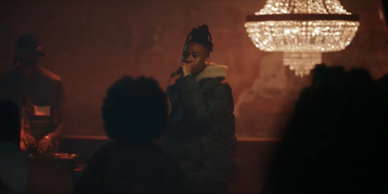 Autumn Beat (2022) movie still: medium — A dimly lit indoor space with a large ornate crystal chandelier hanging above subjects. A man in a puffer…