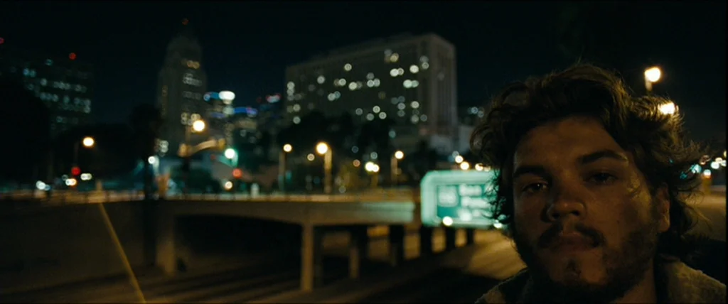 Into the Wild (2007) movie still: closeup — A city highway at night with blurred skyscraper lights visible in the distance. A man stands in the…
