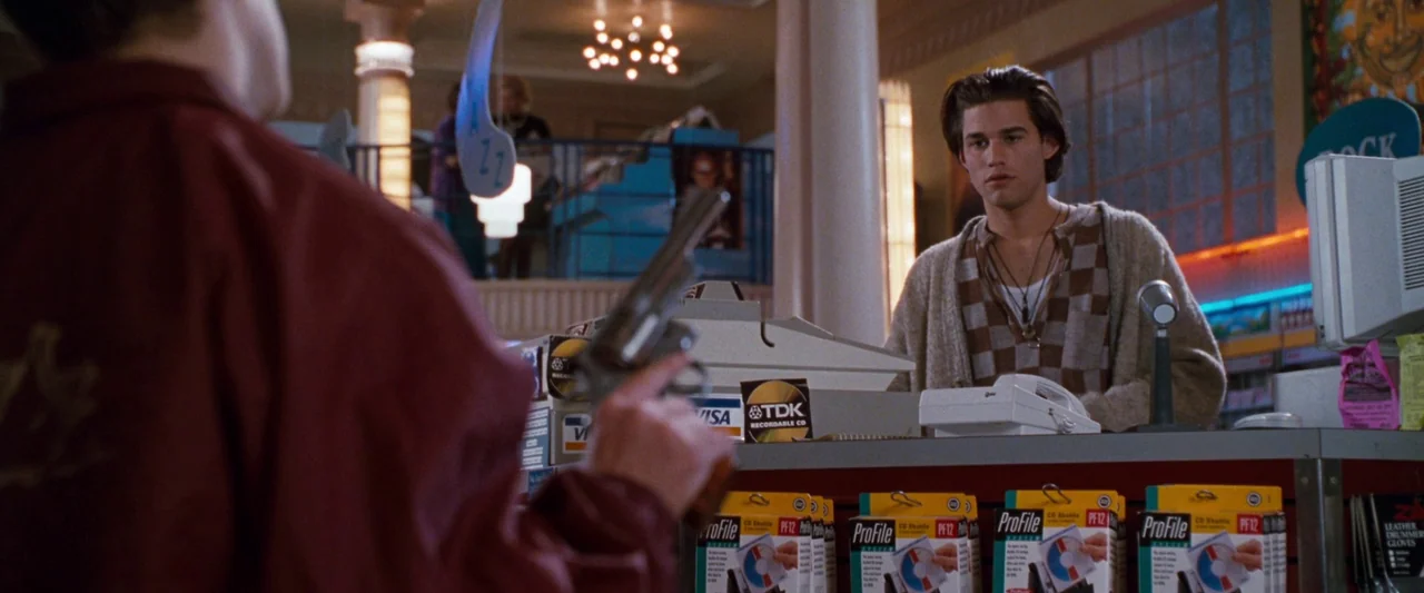 Empire Records (1995) movie still: medium — A 1990s retail electronics store counter is lined with boxes of recordable CDs. A young man stands…
