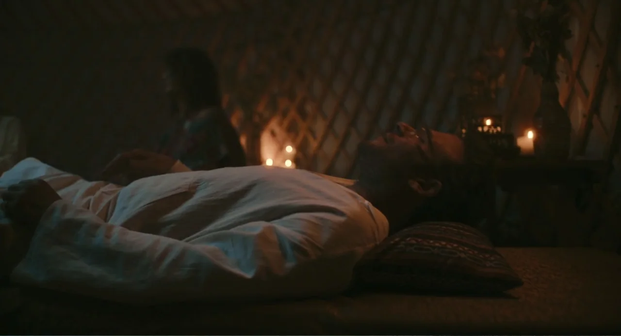 Good Fortune (2025) movie still: medium — A dimly lit interior of a yurt features a wooden lattice wall structure. A man rests on a mattress while a…