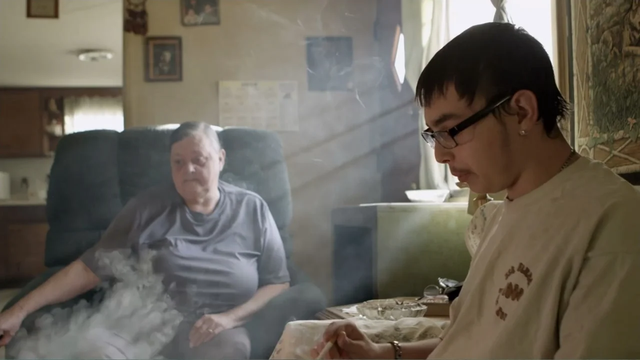 Rich Hill (2014) movie still: medium — A cluttered residential room contains a recliner and table with a glass ashtray. A young man sits in the…