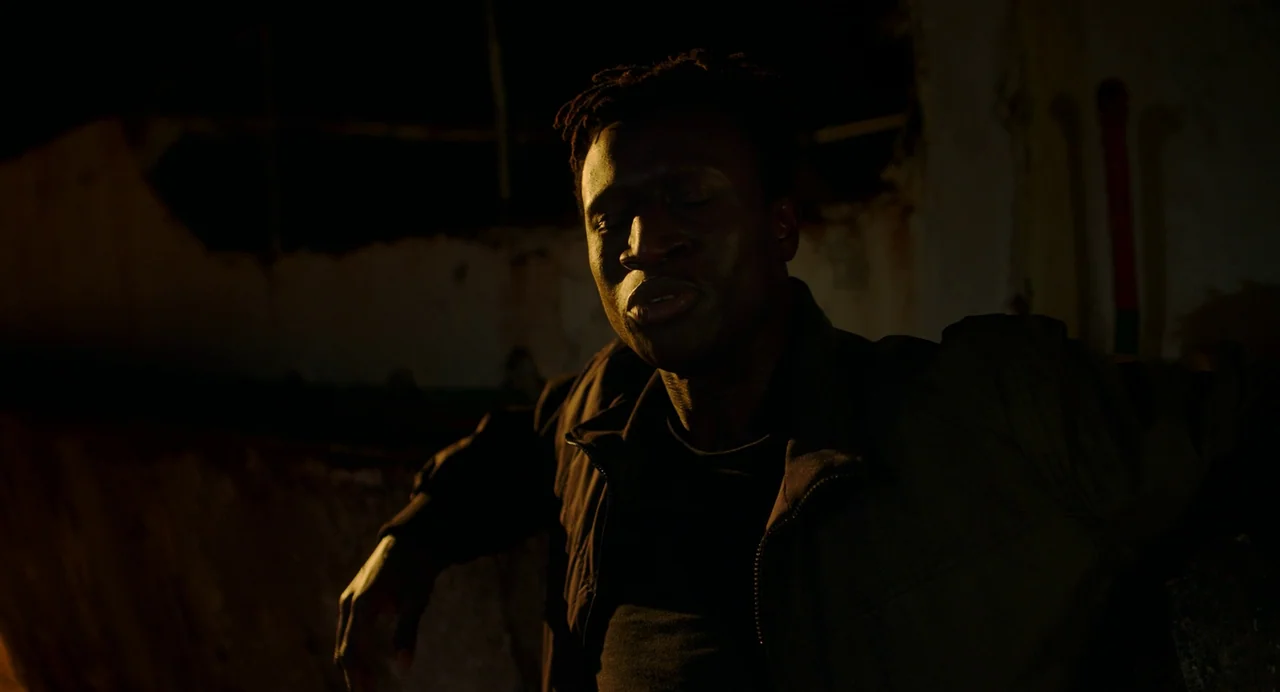 Crimes of the Future (2022) movie still: medium — A dark, decaying industrial space with rough, aged walls in the background. A man sits in low-key…